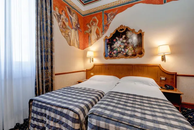 A hotel room with two single beds under a fresco-painted wall, a gilded floral painting above the wooden headboard, and wall lamps on either side.