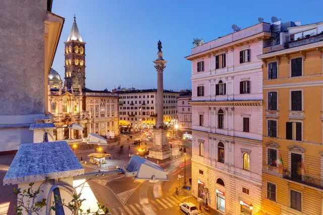 View from an elevated balcony over Piazza Santa Maria Maggiore at dusk, showing the basilica's bell tower, a tall column topped with a statue, and surrounding historic buildings.