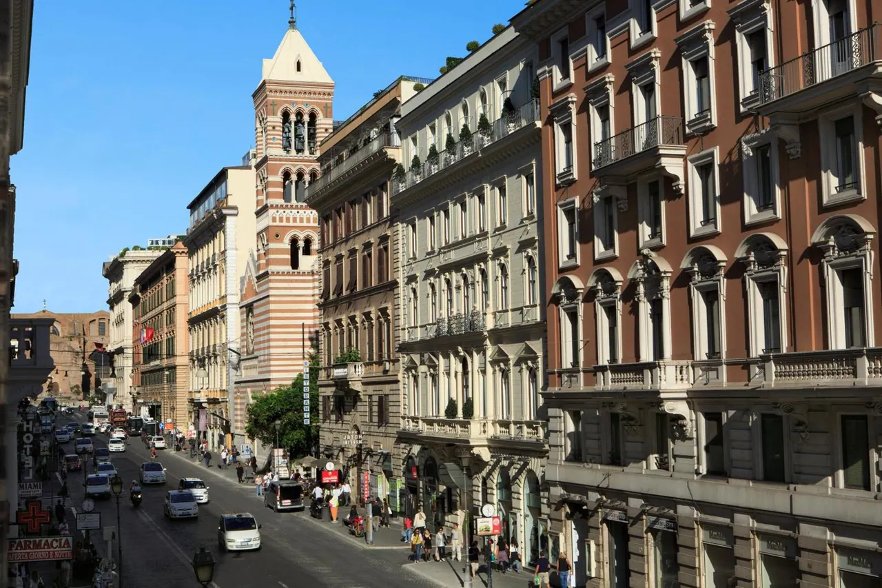 A view down a busy city street lined with old multi-storey buildings, a brick bell tower, and parked and moving cars.
