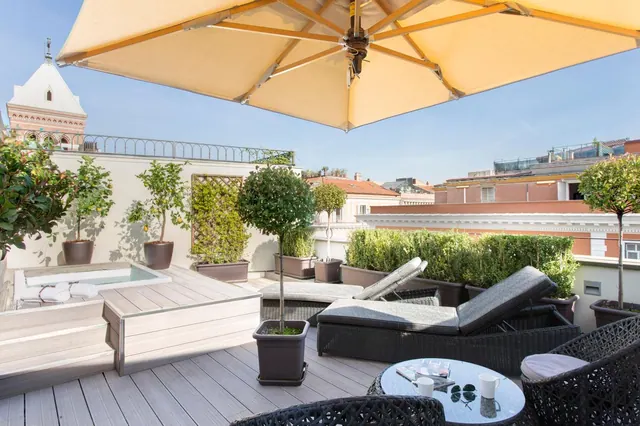 A rooftop terrace with a large parasol, sun loungers, a small sunken jacuzzi, potted trees and a view over neighbouring rooftops and a church tower.