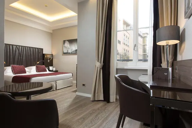 A staged hotel bedroom with a double bed, a desk with a lamp and a round table, and a window looking onto buildings across the street.