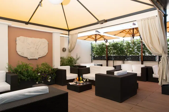 A covered rooftop terrace with wicker lounge seating, white cushions, parasols, drapes and a carved stone relief on the wall.