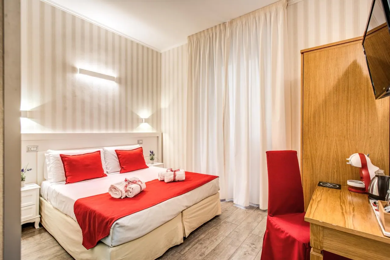 A double bed with red cushions, a red runner and folded towels sits in a bedroom with striped wallpaper, a wooden wardrobe, a desk with a coffee machine and a wall-mounted television.