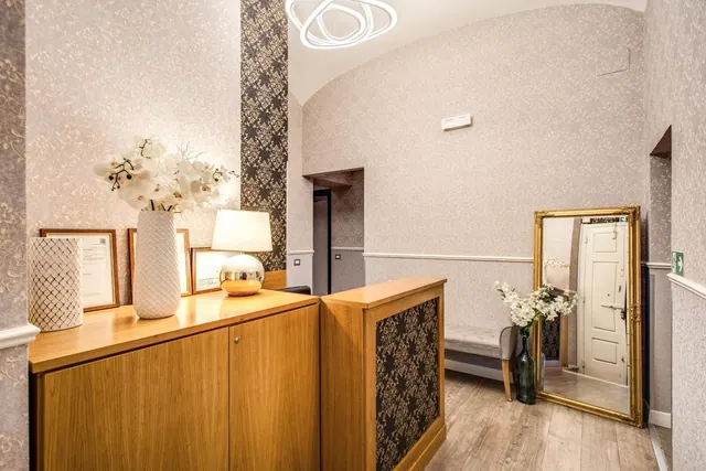 A hotel reception area with a wooden counter topped with a white orchid arrangement, a lamp and framed certificates, beside a gold-framed mirror and an upholstered bench.