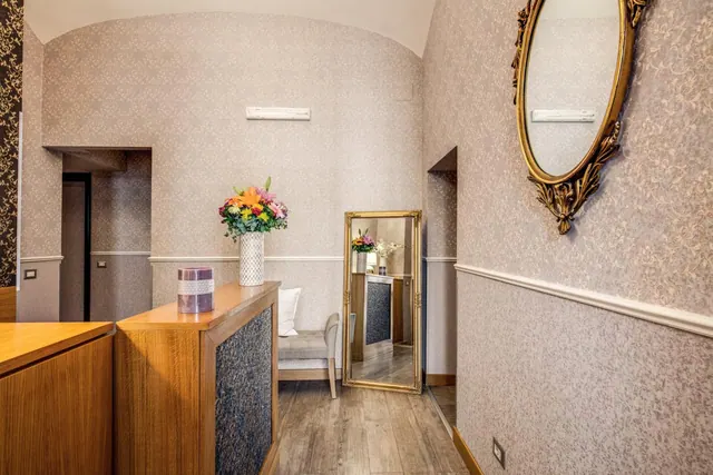 A hotel lobby with a wooden reception desk, a vase of flowers, a gilt-framed mirror and a small upholstered bench against patterned wallpaper.