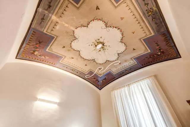 A vaulted ceiling with painted floral and ornamental designs is shown above white walls, a wall light, and a window with white curtains.