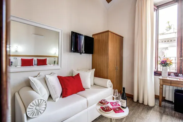 A living area with a white sofa and red cushions, a wall-mounted television, a wooden wardrobe, and a small table with two glasses of wine and two pastries beside a window looking onto a neighbouring building.