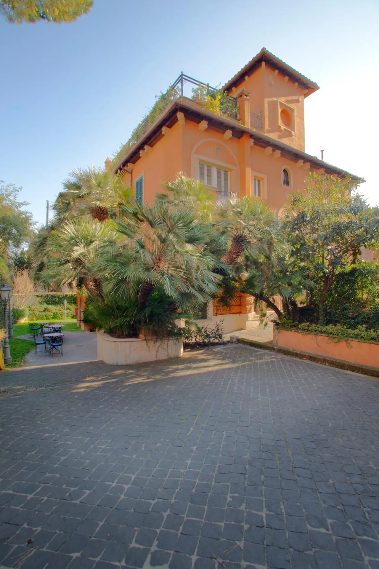 Exterior of an orange villa with a rooftop tower and palm trees, viewed across a cobblestone driveway, with a small outdoor seating area on the left.