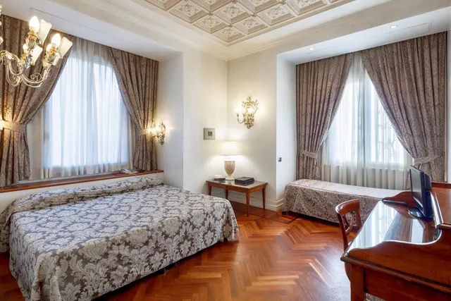 A bedroom with a double bed, a single bed, a crystal chandelier, a wooden writing desk with a television, and a herringbone parquet floor.