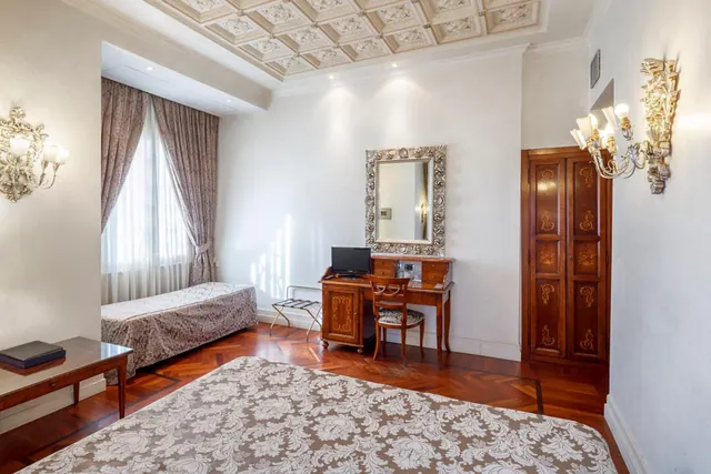 A bedroom with a large double bed in the foreground, a single bed by the window, a desk with a television and chair, and a wooden wardrobe beneath a coffered ceiling.