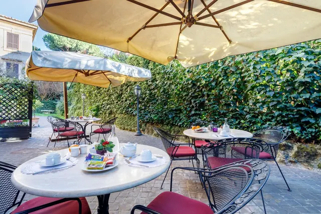 A breakfast table set with cups, a teapot, snacks and juice sits on a paved terrace under cream umbrellas, with an ivy-covered wall behind.