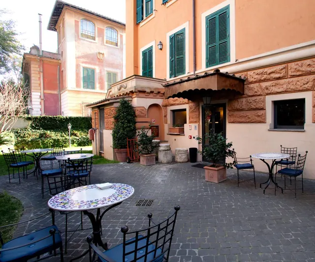 An outdoor courtyard terrace with mosaic-topped tables and wrought iron chairs with blue cushions, in front of an ochre building with green shutters and a covered entrance.