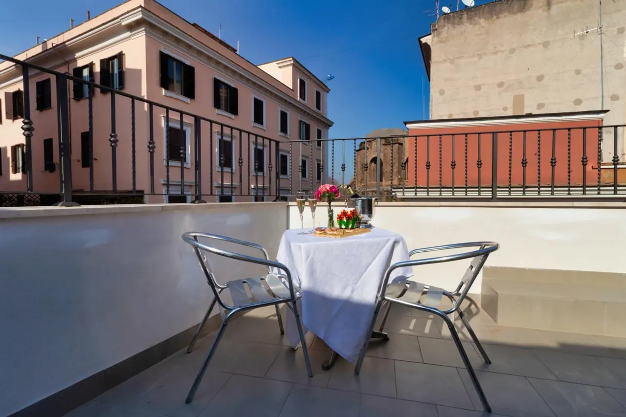 A small table with a white tablecloth, champagne glasses and pink flowers sits on a tiled balcony facing neighbouring buildings under a blue sky.
