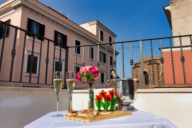 A set table on a balcony holds two champagne flutes, a bottle in an ice bucket, a vase of roses and a board of canapés, with a pink building and an old brick structure behind.