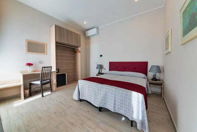 A bedroom with a double bed with a red headboard and runner, a desk with a chair, a wardrobe, a small fridge and a wall-mounted air conditioner.