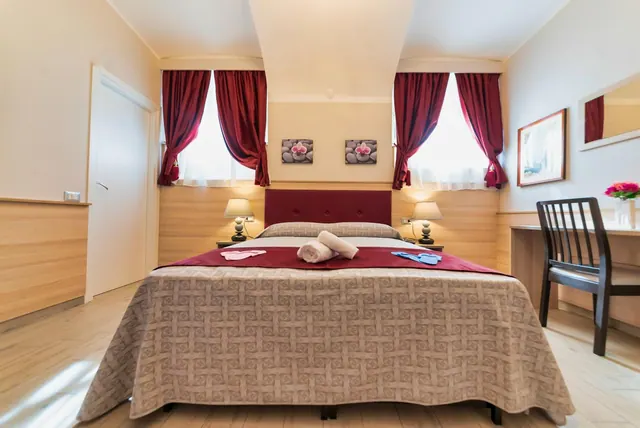 A double bed with a red headboard and red runner sits between two bedside lamps, with red curtains at the windows and a desk and chair on the right.