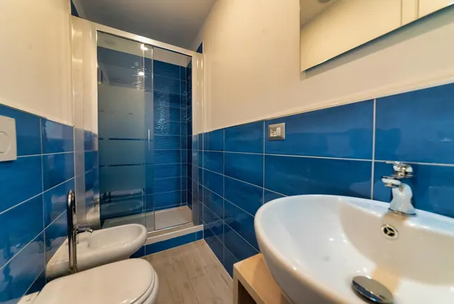 A bathroom with blue wall tiles, a glass-enclosed shower, a bidet and toilet, and a wall-mounted oval basin with a chrome tap.
