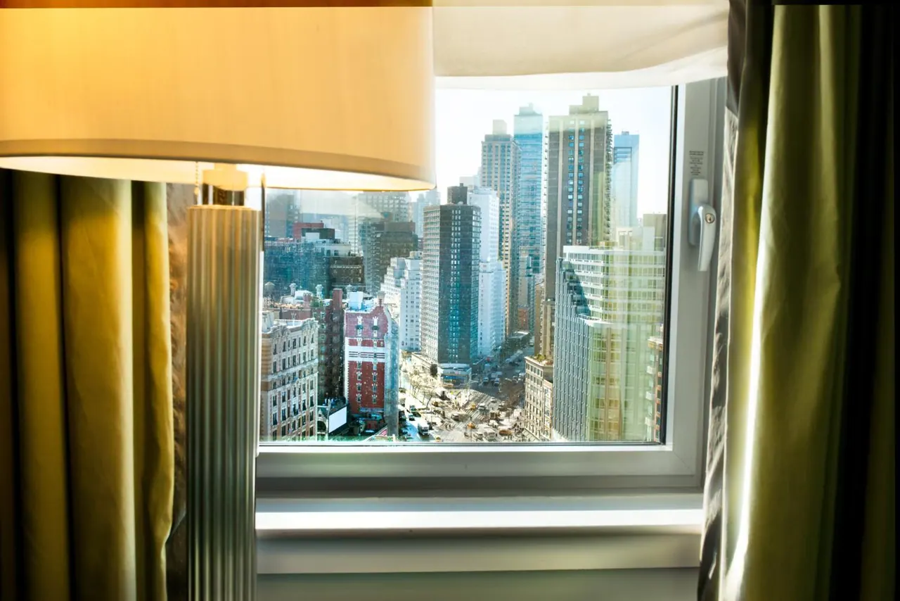 A hotel room window looks out over a city street lined with tall buildings, with a lamp and green curtains in the foreground.