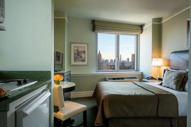 A hotel bedroom with a double bed, a small kitchenette and a window looking out over the Manhattan skyline, including the Empire State Building.