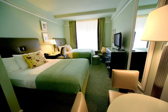 A bedroom with two beds dressed in green bedding, a flat-screen TV on a dresser, an armchair and ottoman, and sheer white curtains over the window.