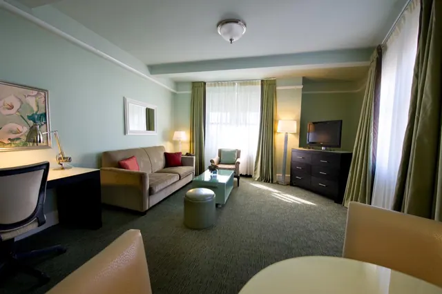 A hotel suite living area with a sofa, armchair, glass coffee table, ottoman, desk with a lamp, and a flat-screen television on a dresser.