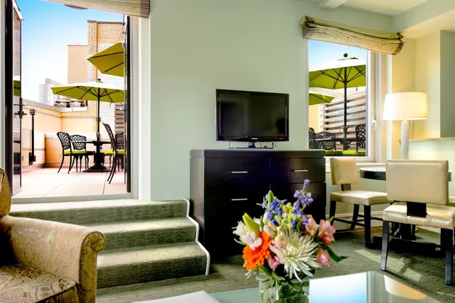 A living area with a flat-screen television on a dark dresser, a short staircase, an armchair, and open doors onto a terrace with green umbrellas and tables.