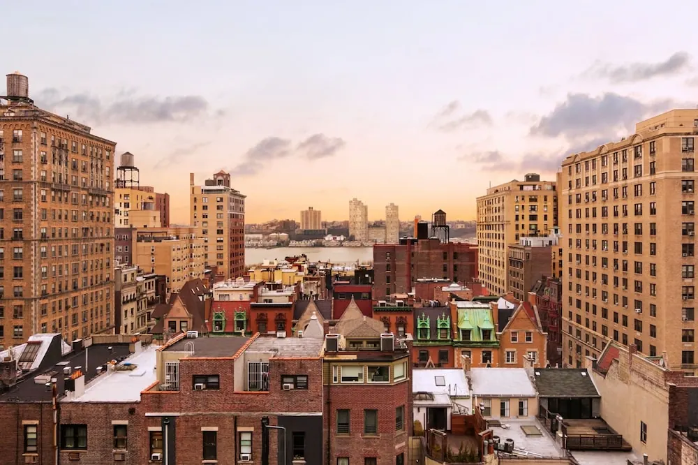 Elevated view over brick rooftops and tall apartment buildings toward a body of water at sunset.