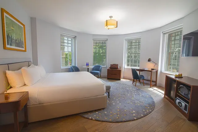 A bedroom with a king bed, blue armchairs, a writing desk, a round rug, a wall-mounted television and windows looking out onto buildings.