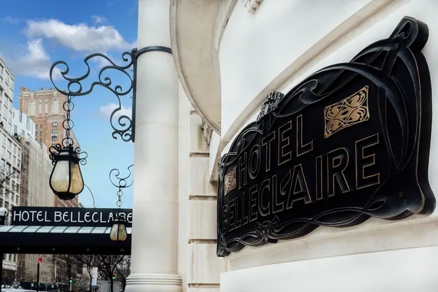 An ornate black and gold Hotel Belleclaire sign is mounted on a white stone facade, with a wrought iron lantern and the hotel awning visible beside it.