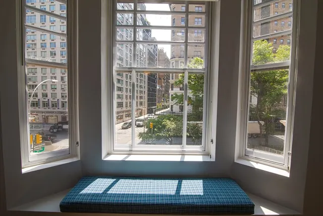 A bay window with a blue plaid cushioned window seat looks out onto a city street lined with apartment buildings and trees.
