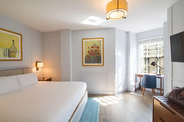 A bedroom with a made king bed, framed artworks, a wall-mounted television, and a desk by a window overlooking a brick building.