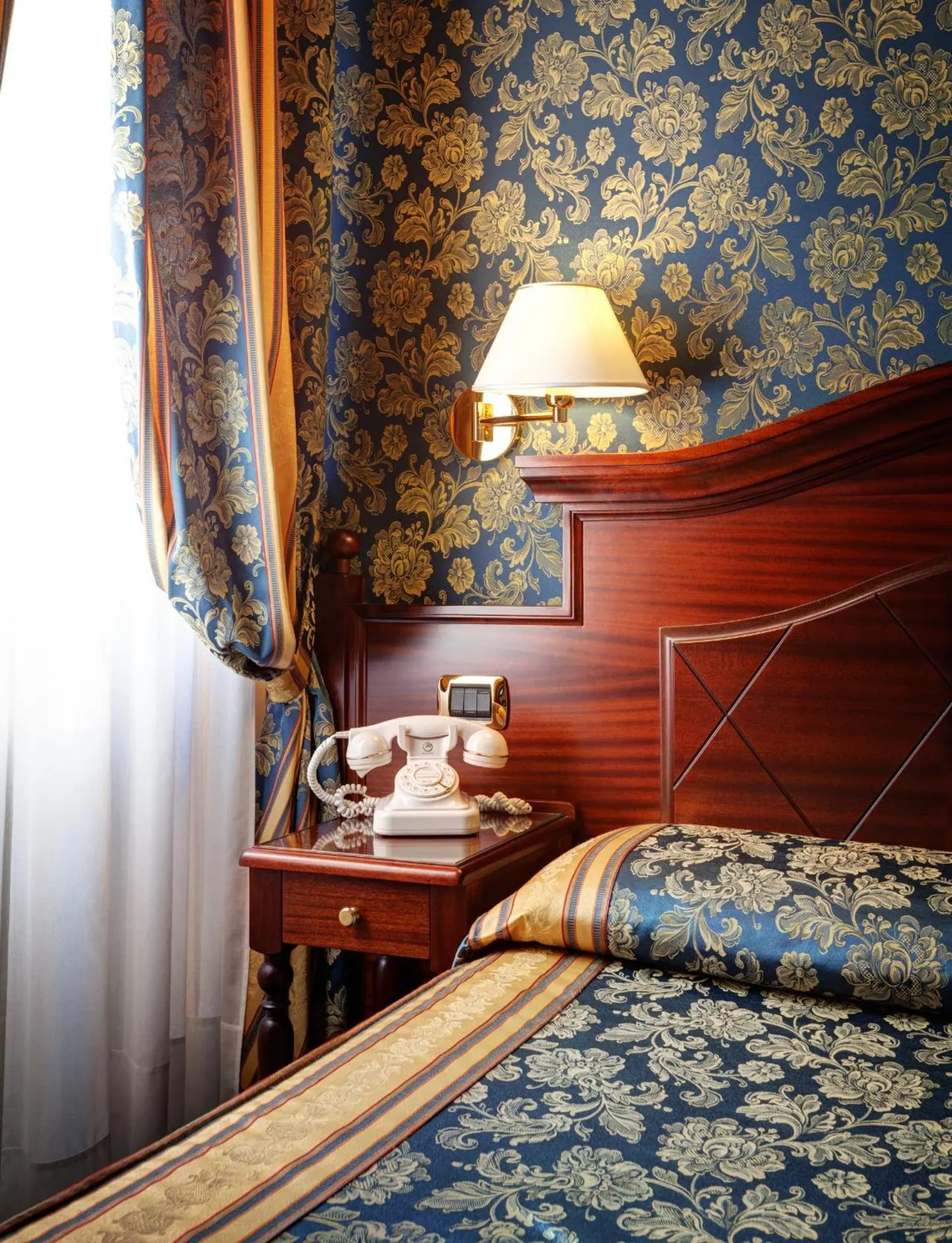 A bedroom with blue and gold patterned wallpaper, a wall lamp, a cream telephone on a wooden nightstand, and a bed with a matching patterned cover beside a curtained window.