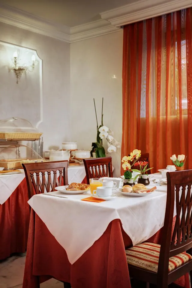 A breakfast room with a set table under white linen, with pastries, orange juice and cups, a buffet along the left wall and orange sheer curtains by the window.