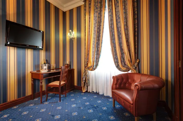 A hotel room with blue and gold striped walls, a wall-mounted television, a wooden writing desk with an ice bucket and glasses, a tufted leather armchair, and sheer white curtains at the window.