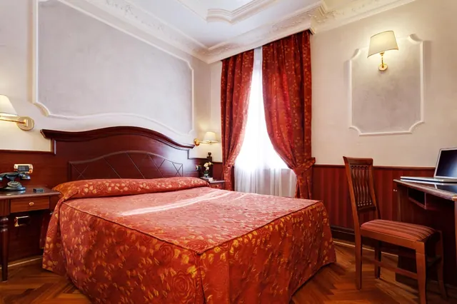 A double bed with a red floral bedspread and wooden headboard sits beside a window with red curtains and a wooden desk with a laptop in a hotel bedroom.