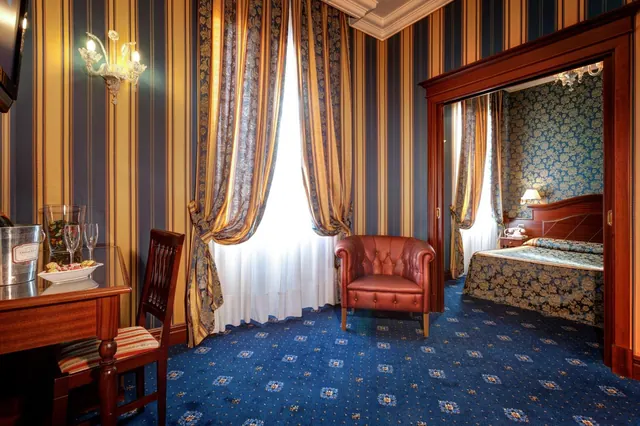 A hotel room with striped wallpaper, a leather armchair and a desk with glasses, with a doorway through to a bed with floral wallpaper.