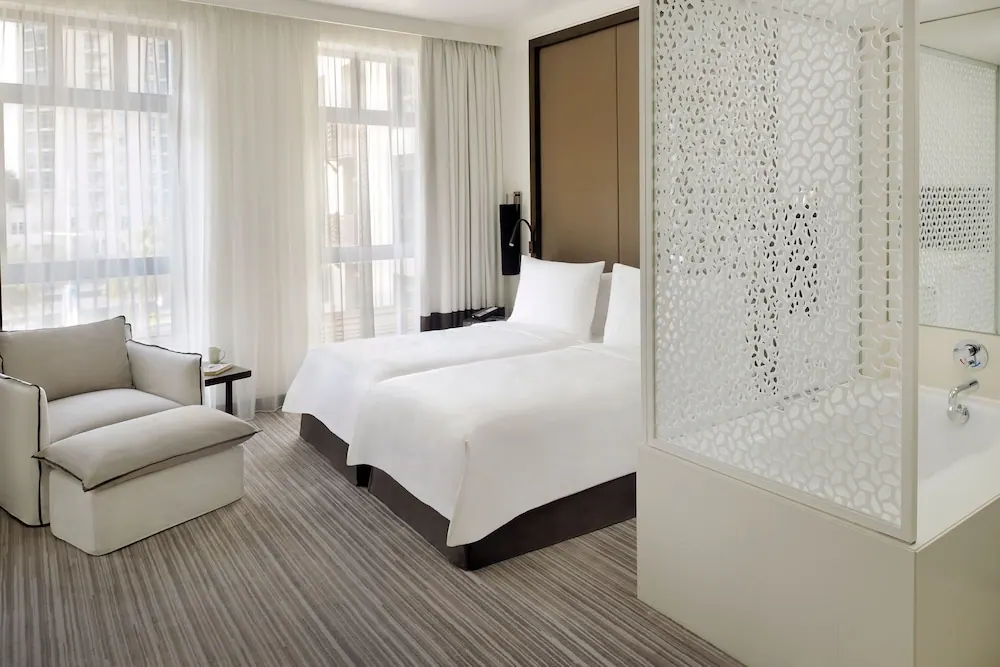 A bedroom with two white twin beds, an armchair and ottoman by sheer-curtained windows, and a bathtub behind a decorative white screen.