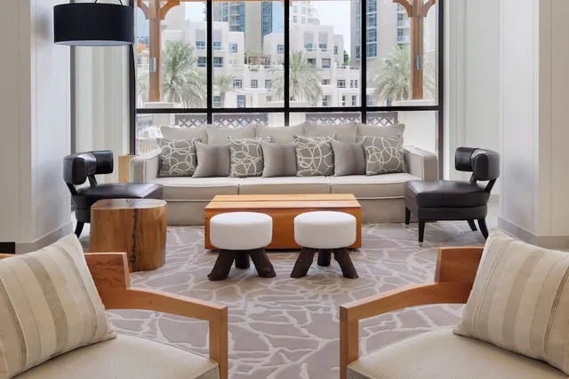 A living area with a grey sofa, dark leather armchairs, a wooden coffee table and stools, with floor-to-ceiling windows showing low-rise buildings and palm trees outside.