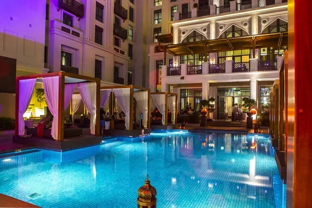 Night view of an illuminated outdoor pool with curtained cabana beds along one side and an arched building with lit balconies behind.