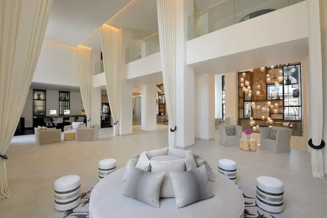 A double-height white lobby with a round cushioned seating arrangement and poufs in the foreground, curtained columns, and a mezzanine with a glass railing behind.