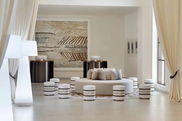 A lobby seating area with a large circular cream sofa, white upholstered stools, a big abstract painting, floor lamps and sheer curtains.