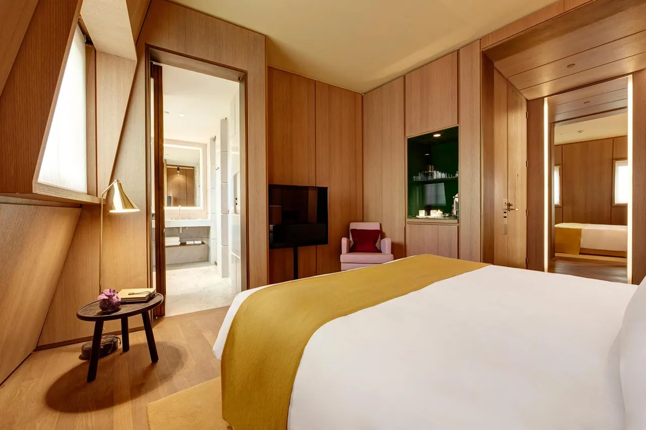 A bedroom with a king bed, wood-panelled walls, a pink armchair, a wall-mounted television and an open doorway to a bathroom with a bathtub.