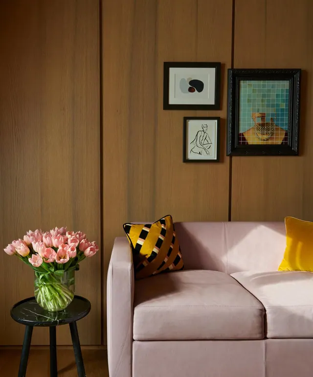 A pink leather sofa with yellow and striped cushions sits beside a wood-panelled wall with framed artworks and a vase of pink tulips on a black side table.