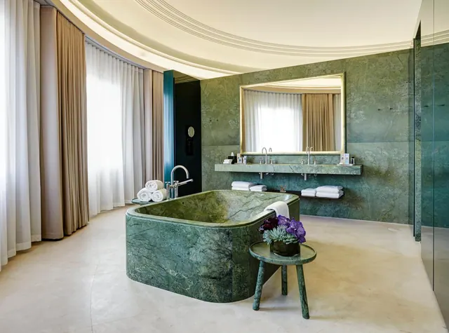 A freestanding green marble bathtub and a double washbasin with a large mirror sit in a bathroom with floor-length curtains and tall windows.