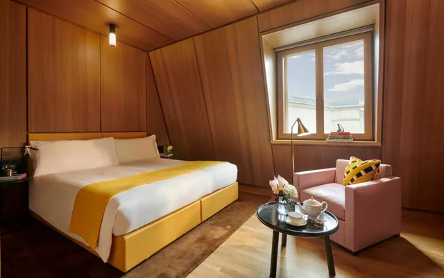 A bedroom with wood-panelled walls, a king bed with a yellow throw, a pink armchair and a round table with a tea set beside a window.