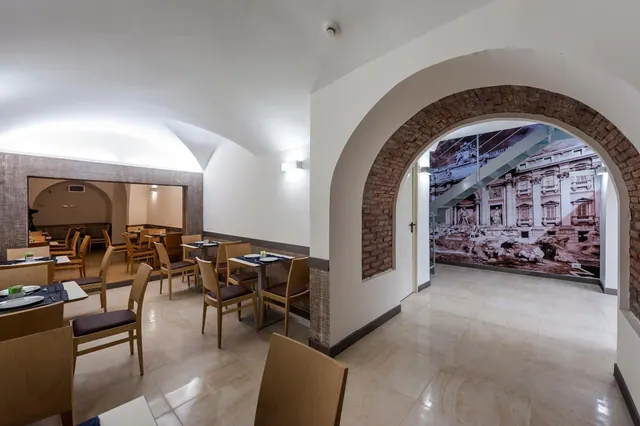 A dining room with wooden chairs and tables, with an arched brick doorway leading to a hallway that has a large photo mural of the Trevi Fountain.