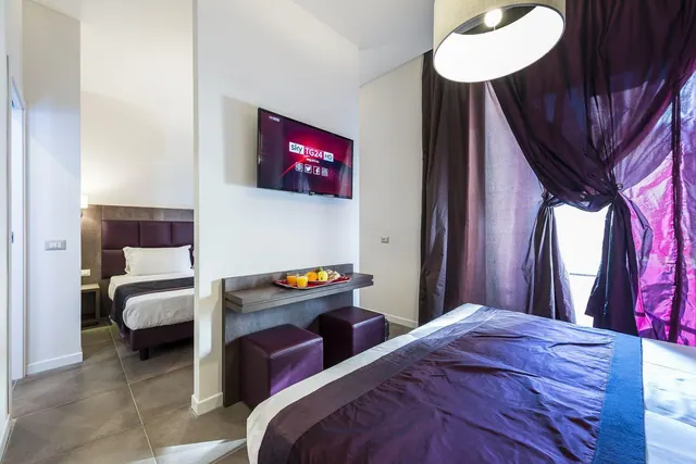 A hotel room with a double bed in purple bedding, purple curtains, a wall-mounted television, a tray of fruit and juice on a wall shelf, and a single bed visible in an adjoining area.