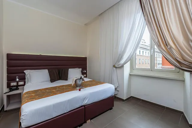 A double bed with a brown leather headboard and a champagne bucket on the bedspread, with a window looking out onto a building across the street.