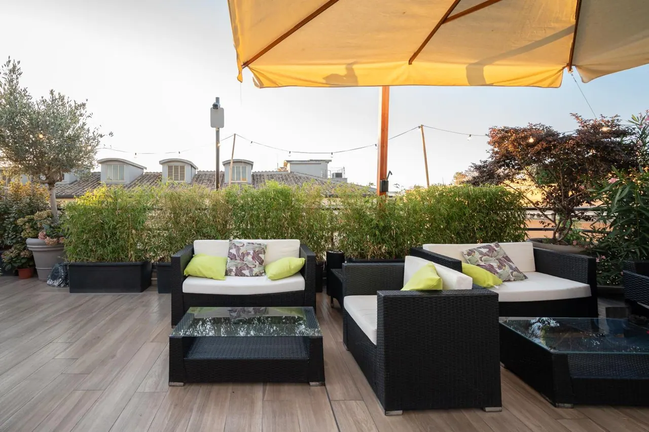 A rooftop terrace with black wicker sofas, cream cushions, glass-topped coffee tables and a yellow parasol, with a bamboo hedge and rooftops behind.