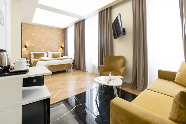 A hotel room with a double bed at the back, a yellow sofa and armchair in the foreground, a wall-mounted television and a kettle on the left counter.
